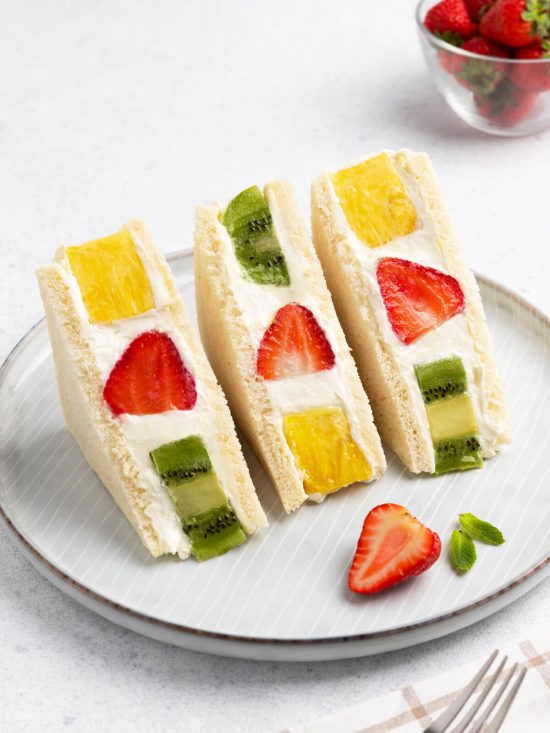 Drei Fruit-Sando-Hälften mit Ananas, Erdbeere und Kiwi in weißer Creme auf einem hellen Teller, daneben eine halbe Erdbeere und Minzblätter