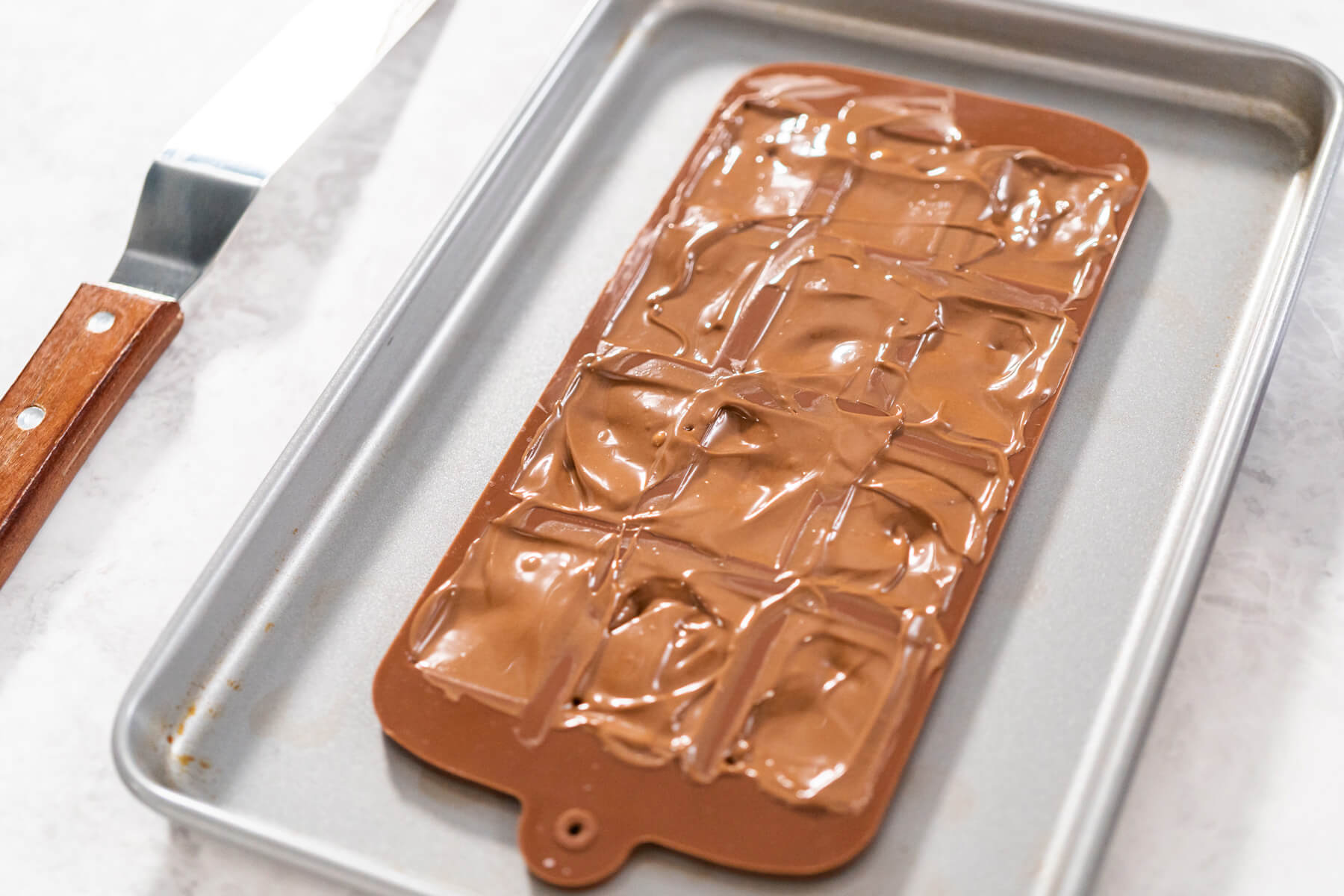 Silikonform mit Mini-Tafel-Mulden voller geschmolzener Vollmilchschokolade auf einem Backblech, daneben ein Winkelspatel