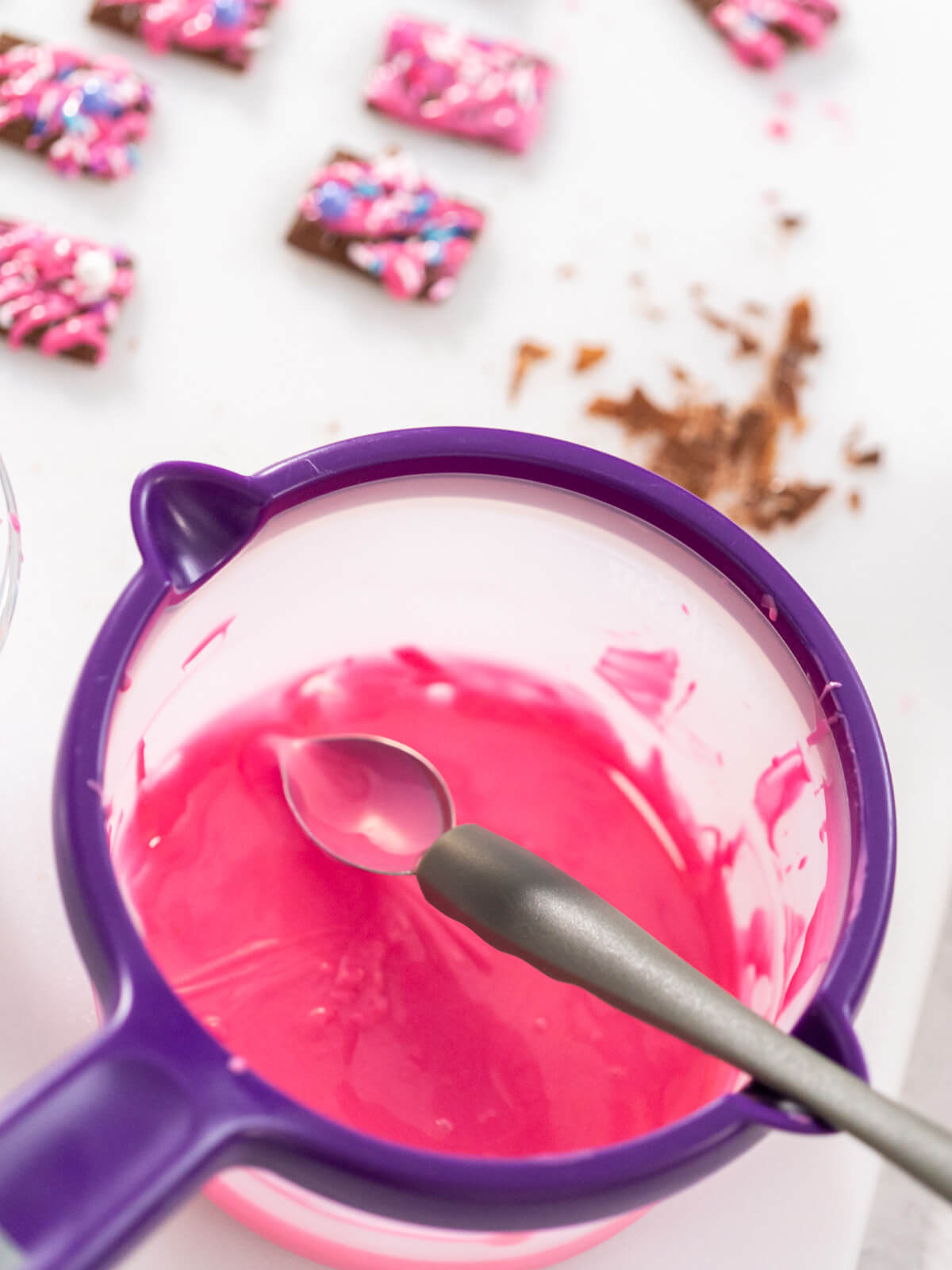 Geschmolzene rosa Schmelzglasur mit Teelöffel in einem violetten Schmelztopf, im Hintergrund fertig verzierte Schoko-Topper