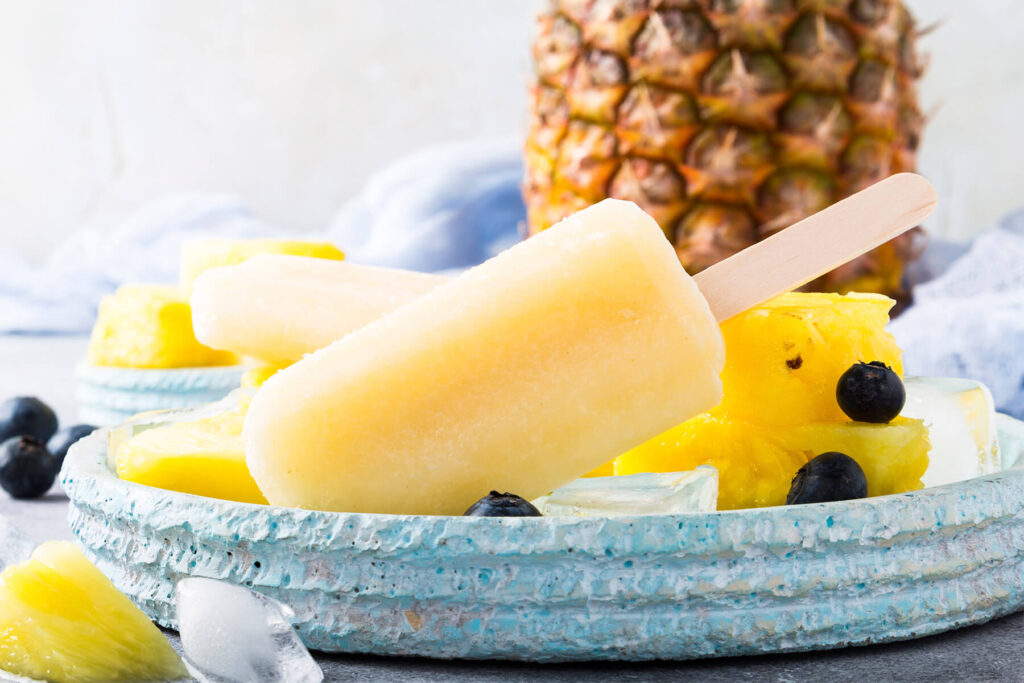 Goldgelbes Ananas-Kokos-Stieleis am Holzstiel mit frischer Ananas, Ananasstücken und Heidelbeeren auf türkiser Keramikplatte