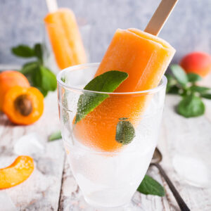Orangefarbenes Aprikosen-Stieleis am Holzstiel mit frischen Aprikosenhälften und Crushed Ice