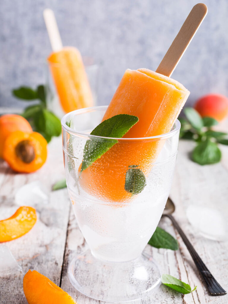 Orangefarbenes Aprikosen-Stieleis am Holzstiel mit frischen Aprikosenhälften und Crushed Ice