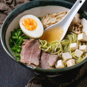 Asiatische Nudelsuppe mit grรผnen Spinat-Ramen, Rindfleisch, Tofu, Enoki-Pilzen und weichem Ei in dunkler Keramikschale
