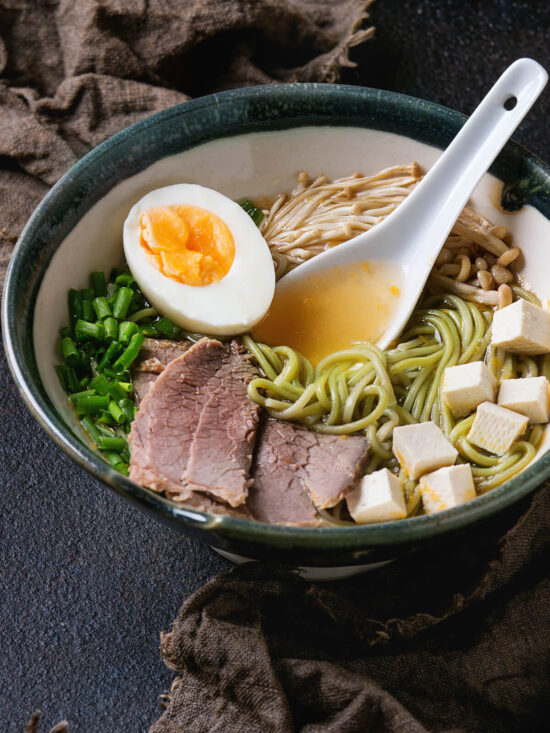 Asiatische Nudelsuppe mit grünen Spinat-Ramen, Rindfleisch, Tofu, Enoki-Pilzen und weichem Ei in dunkler Keramikschale