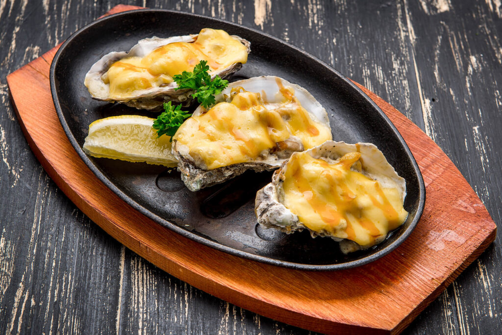 Drei überbackene Austern mit Käse, Zitronenspalte und Petersilie auf gusseiserner Servierplatte