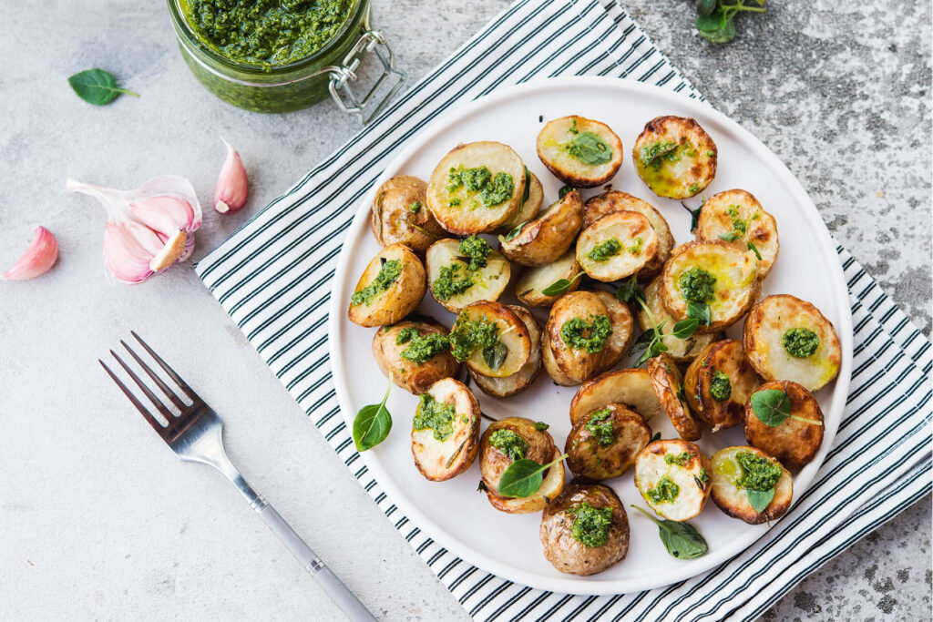 Selbstgemachtes grünes Basilikum-Pesto im Glas neben gerösteten Babykartoffeln und Knoblauch