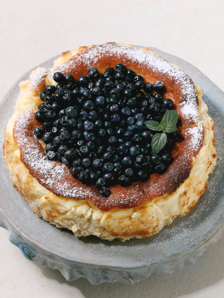 Baskischer burnt Cheesecake aus San Sebastián mit dunkler karamellisierter Oberfläche, frischen Heidelbeeren, Puderzucker und Minze auf Keramik-Tortenständer