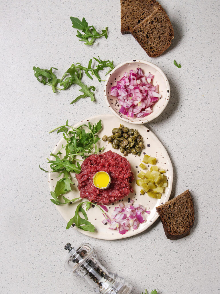 Klassisches Beef Tartare mit Wachtelei in der Mitte, Kapern, Cornichons, roter Zwiebel und Rucola auf Keramikteller
