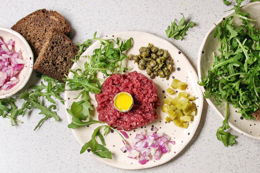 Klassisches Beef Tartare mit Wachtelei in der Mitte, Kapern, Cornichons, roter Zwiebel und Rucola auf Keramikteller
