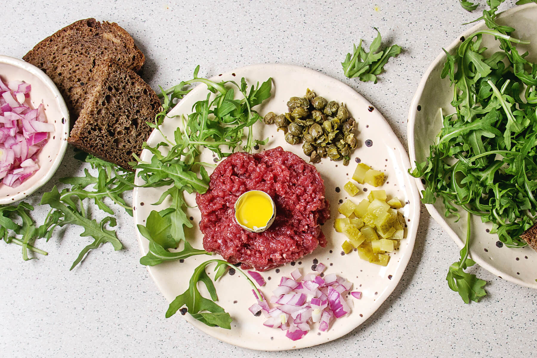 Klassisches Beef Tartare mit Wachtelei in der Mitte, Kapern, Cornichons, roter Zwiebel und Rucola auf Keramikteller