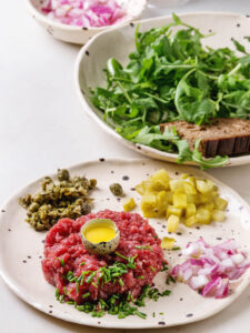 Klassisches Beef Tartare mit Wachtelei in der Mitte, frisch gehackter Schnittlauch, Kapern, gewürfelte Cornichons und rote Zwiebel auf Keramikteller