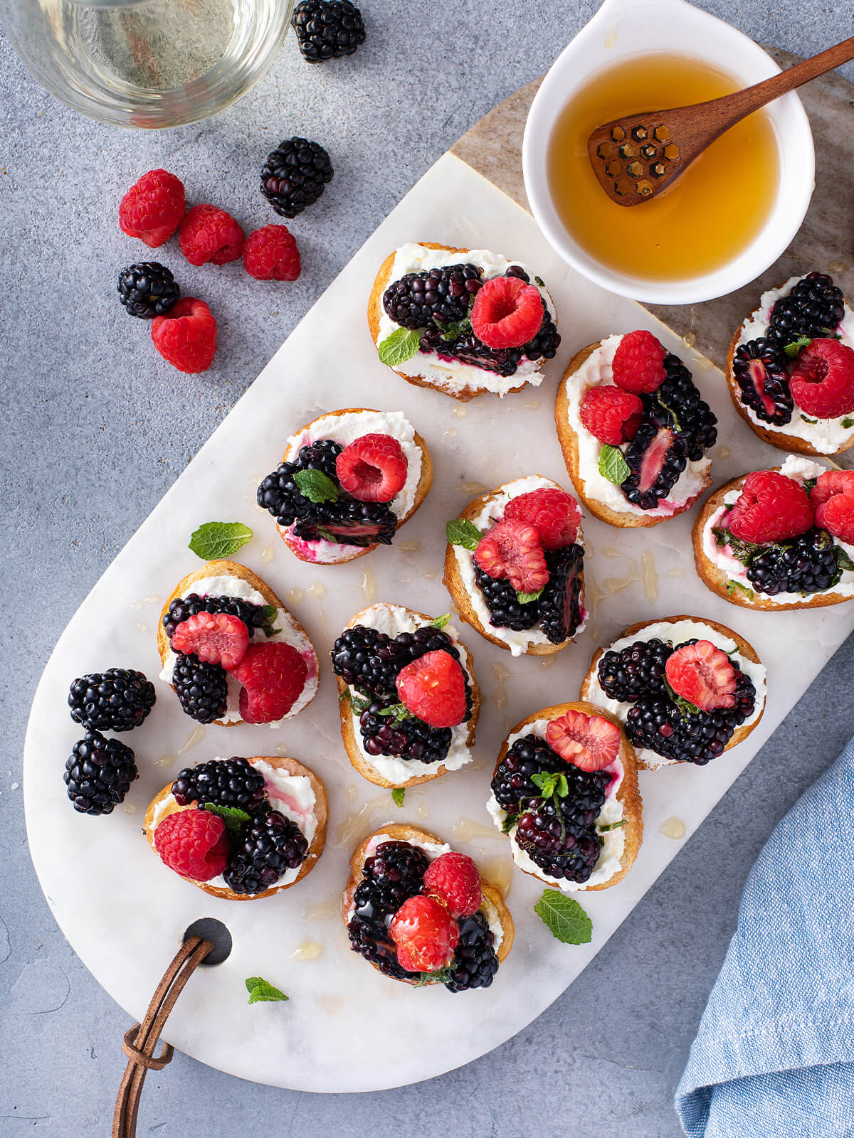 "Beeren-Bruschetta von oben auf einer Marmorplatte angerichtet, mit Brombeeren, Himbeeren, Erdbeeren, Minze und einer Schale Honig"