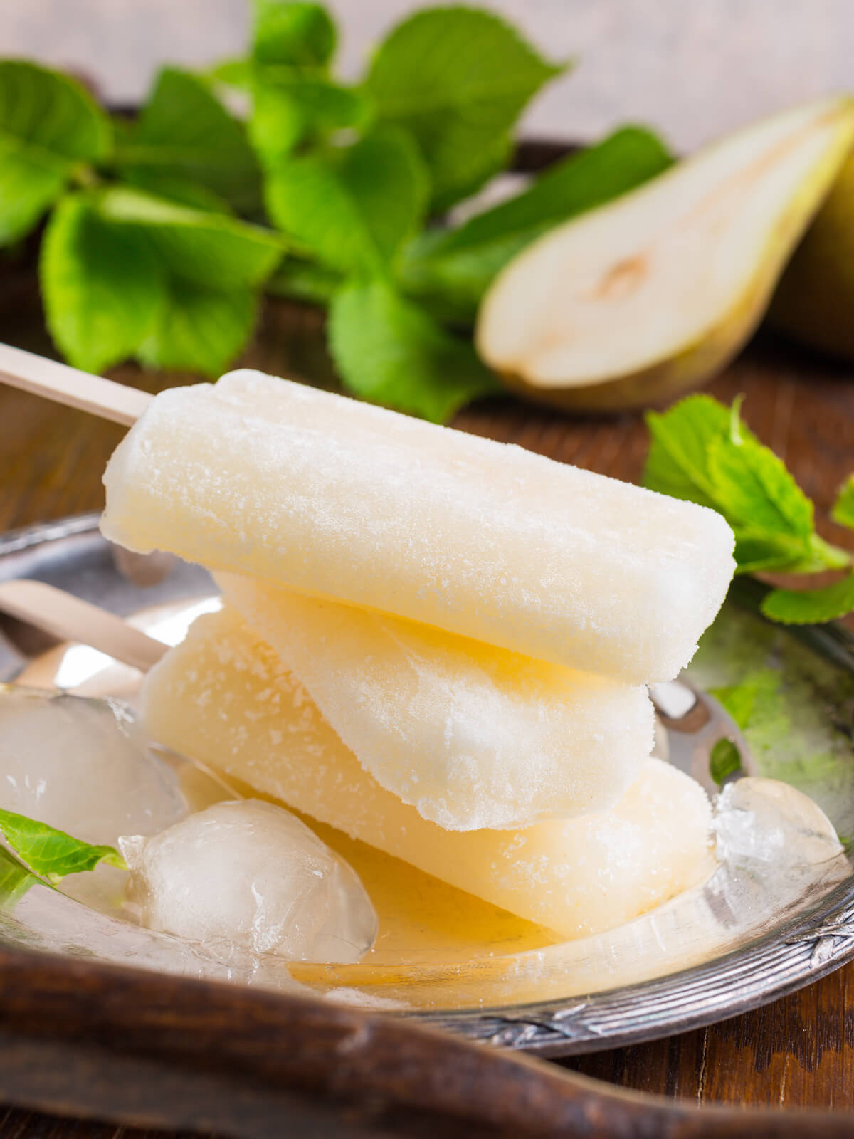 Cremig-weiße Birnen-Sorbet-Stieleis-Stücke auf silbernem Tablett mit Crushed Ice und halber frischer Birne