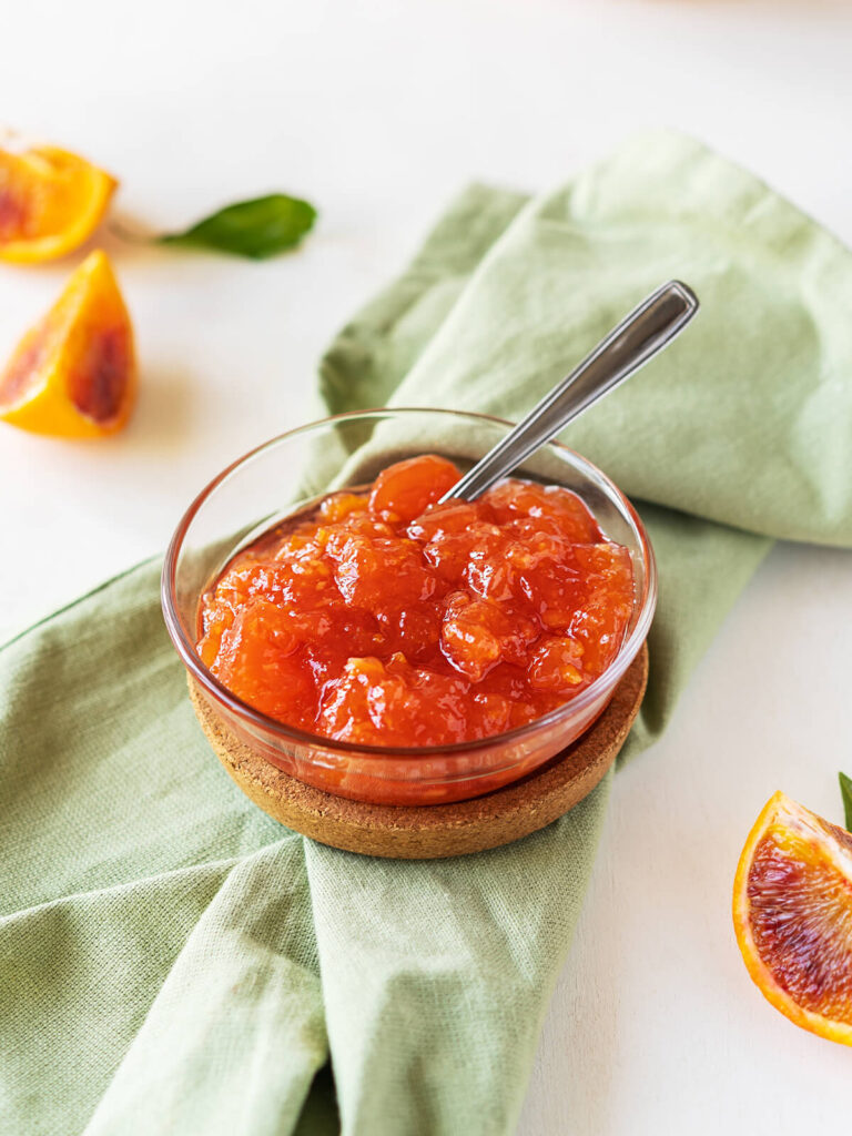 Hausgemachte Blutorangen-Marmelade in einer Glasschale mit Teelöffel, auf grüner Leinenserviette mit halbierten Blutorangen daneben