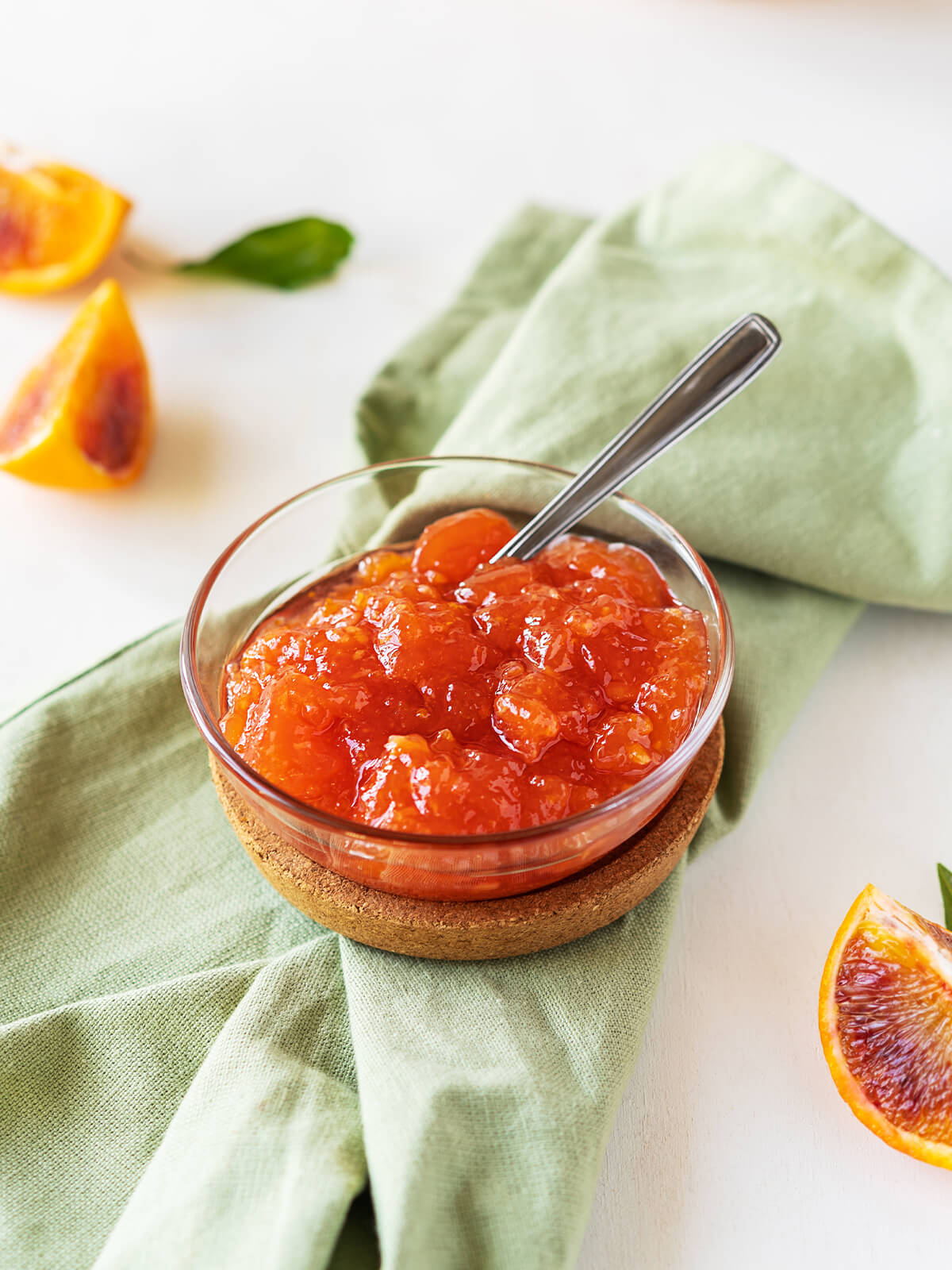 Hausgemachte Blutorangen-Marmelade in einer Glasschale mit Teelöffel, auf grüner Leinenserviette mit halbierten Blutorangen daneben