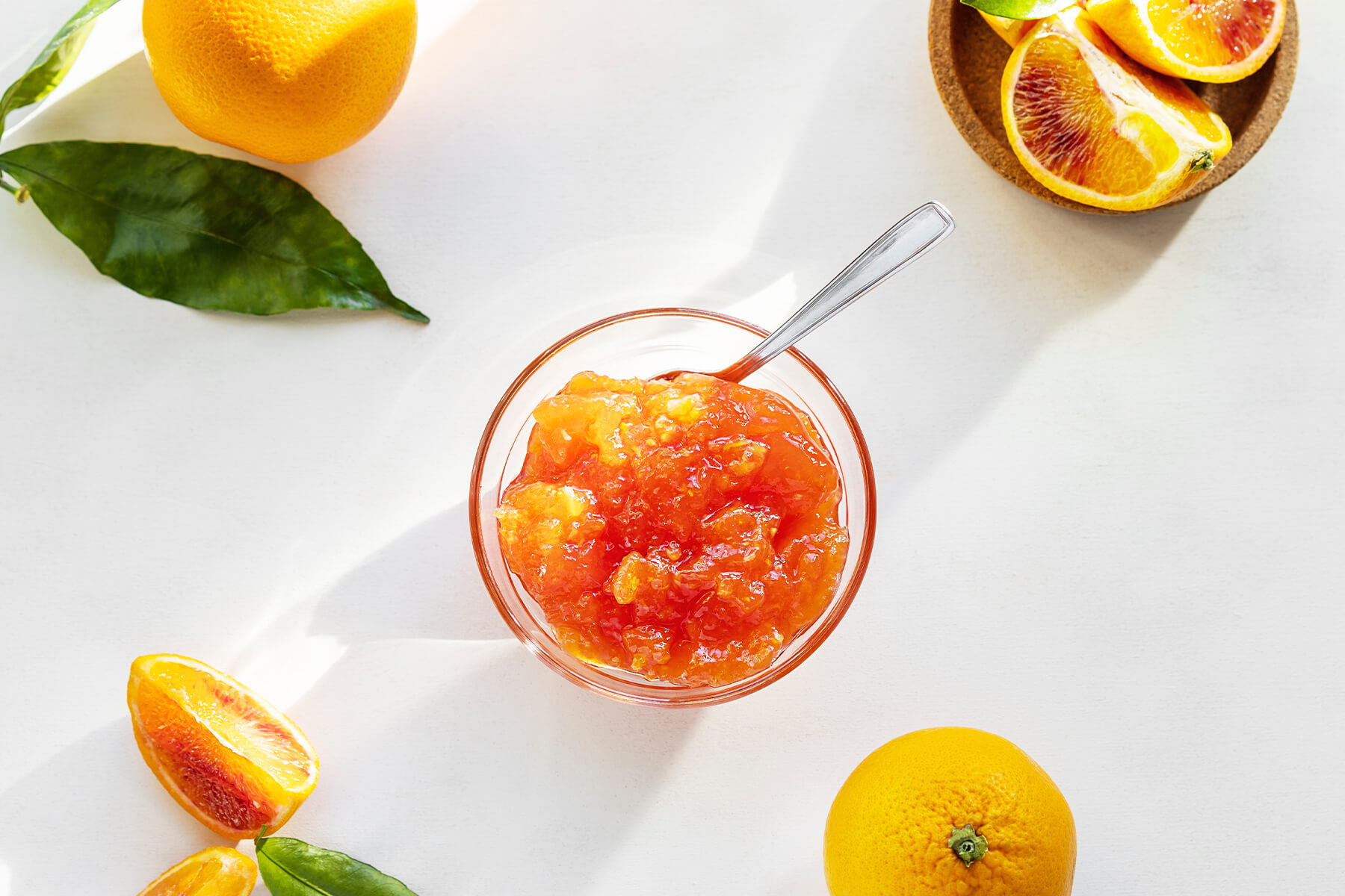 Blutorangen-Marmelade in einer Glasschale mit Löffel, von oben fotografiert, daneben ganze und aufgeschnittene Blutorangen mit rotem Fruchtfleisch