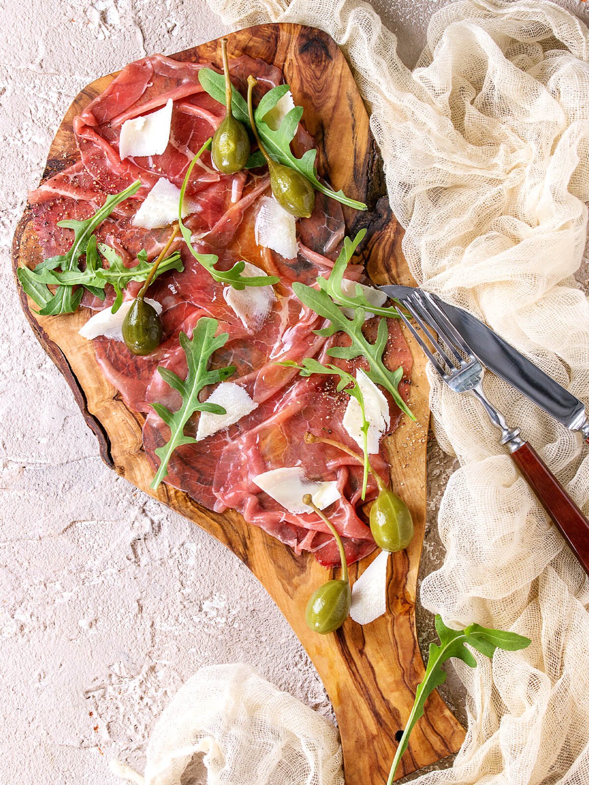 Bresaola-Carpaccio mit Rucola, Parmesan und Kapernäpfeln auf einem Olivenholzbrett, daneben Besteck und ein leichtes Tuch