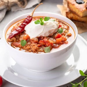 Chili con Carne in weißer Schüssel mit Schmand, Oregano und getrockneter Chilischote