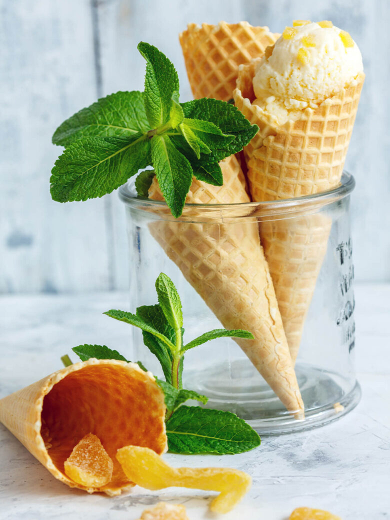 Drei blassgelbe Kugeln Ingwereis in einer Waffeltüte mit kandierten Ingwerstücken und frischer Minze