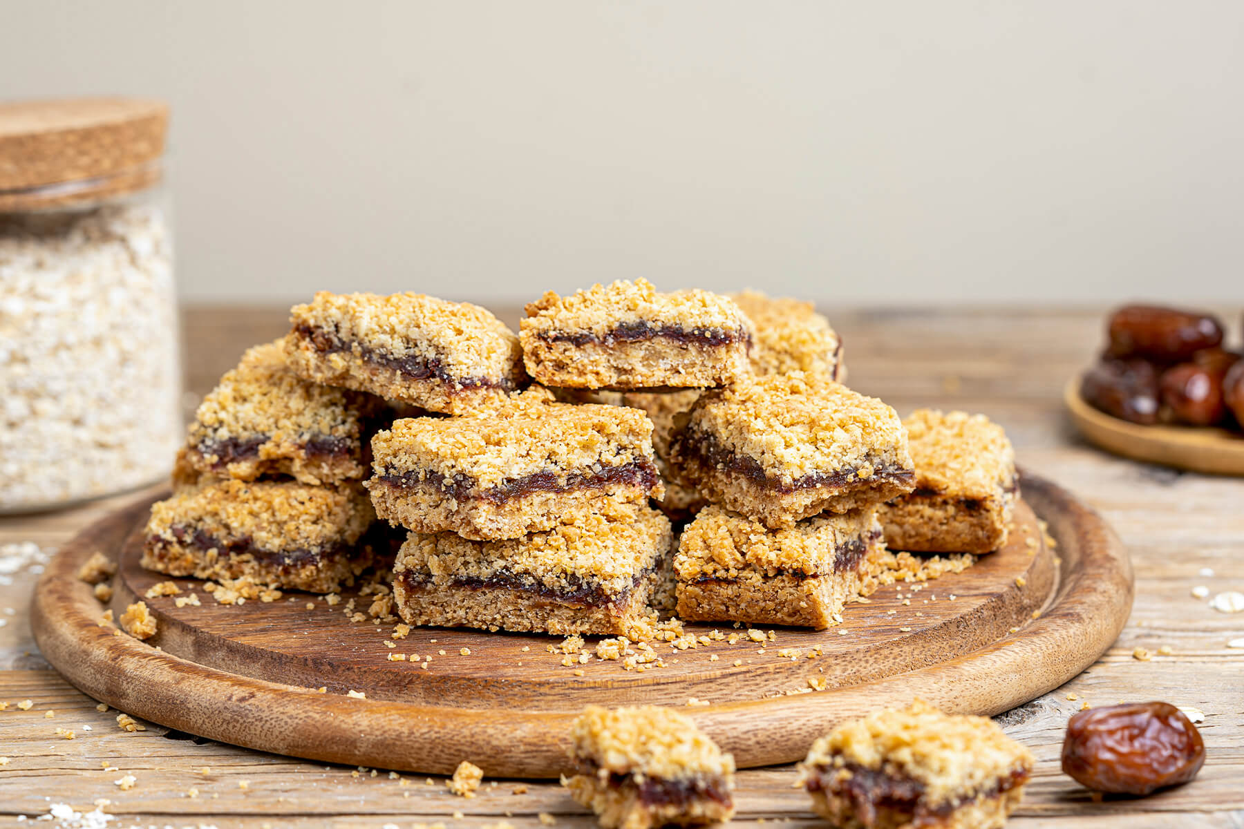 Mehrere kanadische Date Squares auf einem runden Holzbrett mit Datteln und Haferflocken im Hintergrund