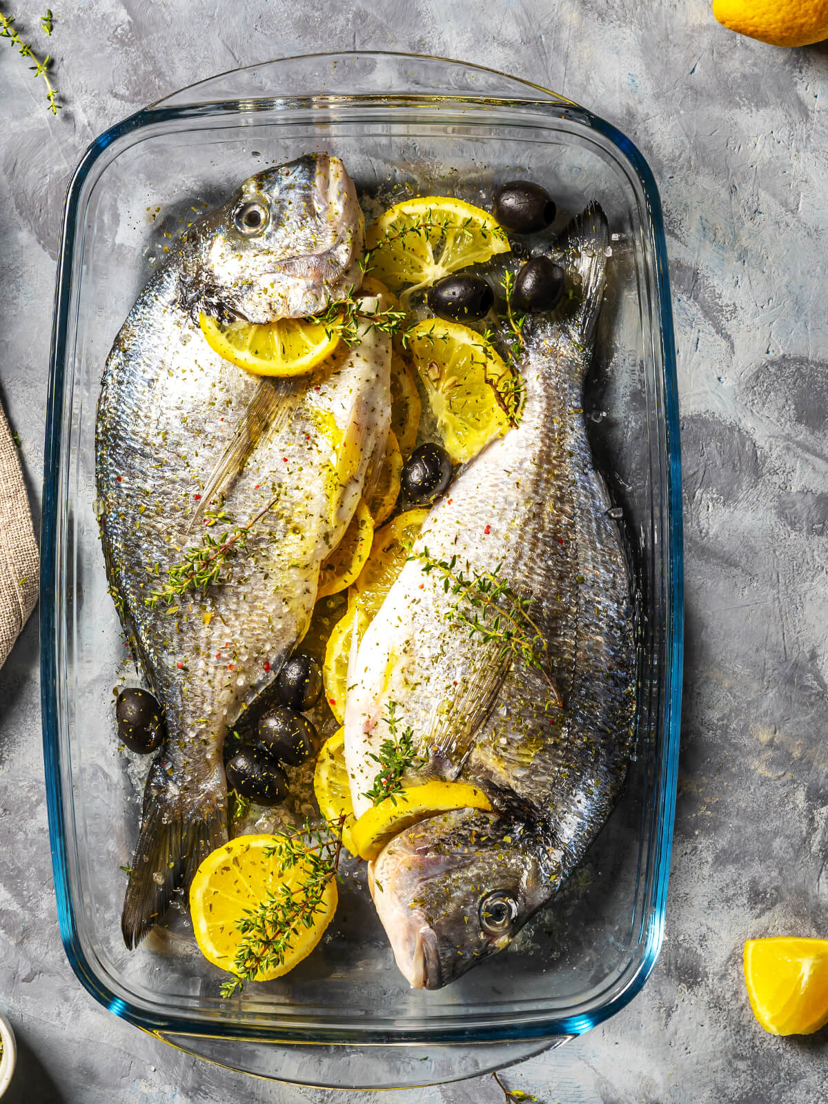 Zwei rohe Dorade-Fische in Glasform mit Zitronenscheiben, schwarzen Oliven und Thymianzweigen bereit zum Backen im Ofen
