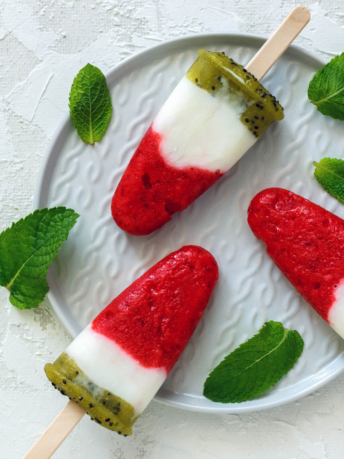 Dreischichtiges Stieleis mit roter Erdbeerschicht, weißer Joghurtmitte und grüner Kiwischicht auf weißem Teller