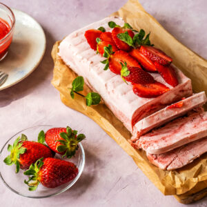 Marmoriertes Erdbeer-Semifreddo in Kastenform mit frischen Erdbeeren, Minze und einem Glasschälchen Erdbeer-Coulis auf rosa Hintergrund