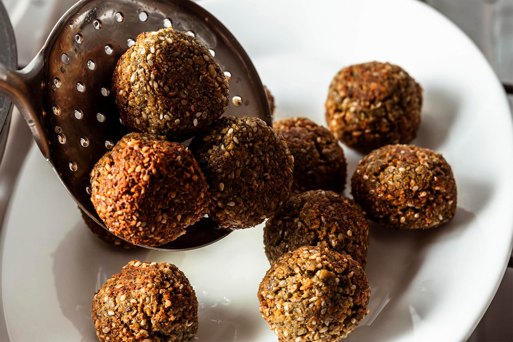 Frittierte Falafel mit Sesamhülle auf einem Schaumlöffel und einem weißen Teller