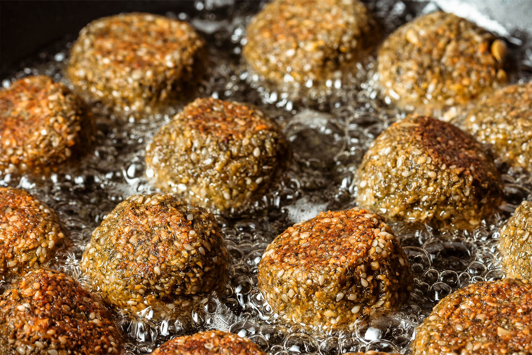 Falafel-Bällchen mit Sesamhülle in flachem heißem Öl, unten goldbraun, oben noch hell