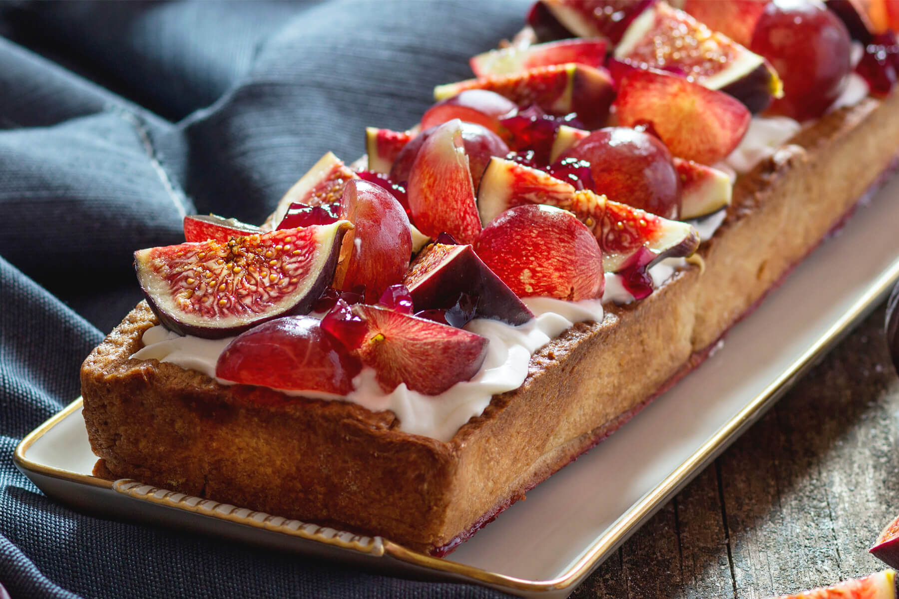 Nahaufnahme einer Feigen-Trauben-Tarte, zwischen den Fruchtstücken sitzen glänzende Tupfen roten Gelees auf der weißen Creme