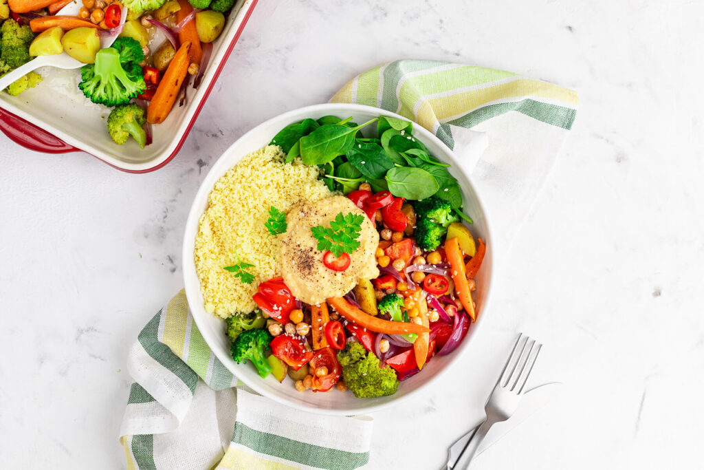 Fertige Gemüse-Bowl mit Hummus, Couscous, Babyspinat und Ofengemüse neben der Auflaufform mit dem restlichen Gemüse