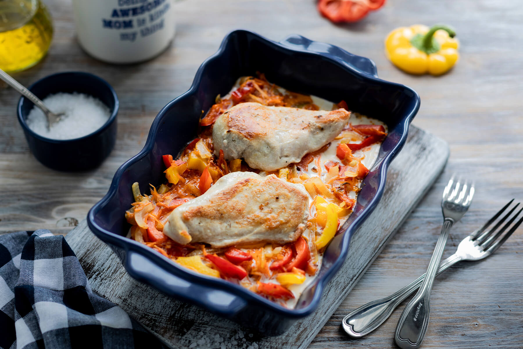 Hähnchenfilet mit buntem Paprika und geriebenen Möhren in dunkelblauer Keramikform, daneben Gabeln und Salz