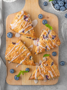 Vier Heidelbeer-Scones mit Zitronenglasur-Streifen auf einem Holzbrett, dazu frische Heidelbeeren