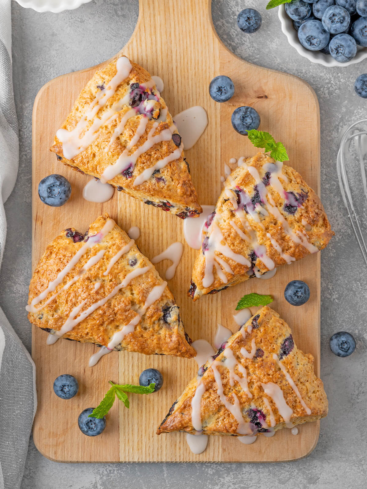 Vier Heidelbeer-Scones mit Zitronenglasur-Streifen auf einem Holzbrett, dazu frische Heidelbeeren
