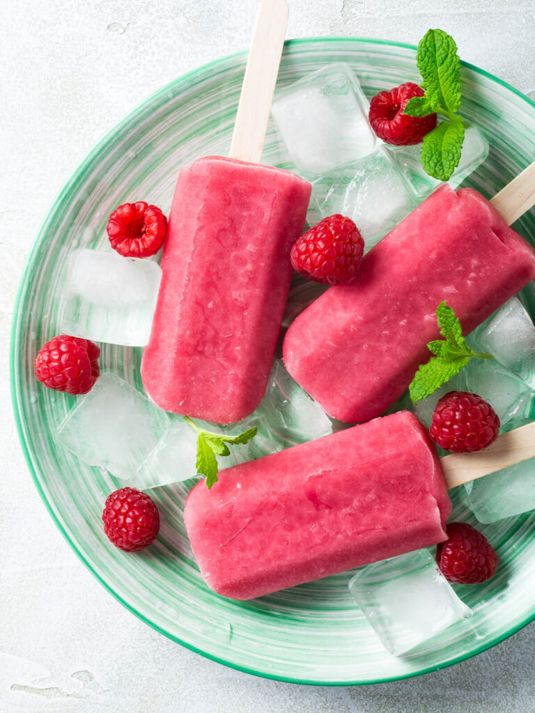 Leuchtend pinke Himbeer-Joghurt-Stieleis-Stücke auf einem Tablett mit Crushed Ice, frischen Himbeeren, gefrorenen Beeren und Minzblättern