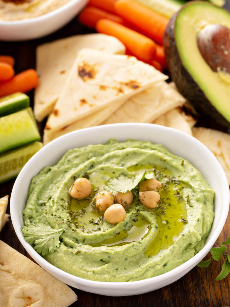 Cremiger grüner Hummus mit Avocado, Koriander und Kichererbsen in weißer Schale mit Karotten, Pita und halbierter Avocado