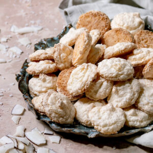 Kokos-Baiser mit heller Oberfläche und goldbrauner Unterseite auf blaugrünem Keramikteller, daneben grobe Kokoschips auf rosa Untergrund