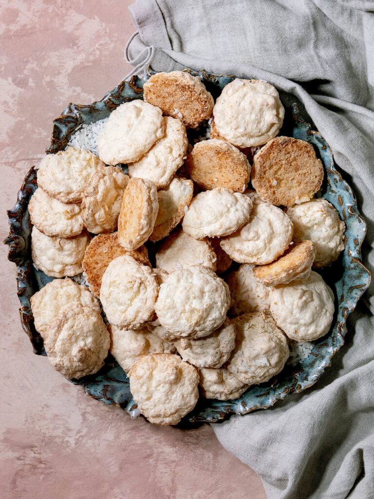 Kokos-Baiser mit heller feinrissiger Oberfläche auf blaugrünem Keramikteller, dazwischen umgedrehte Baiser mit goldbrauner Unterseite, von oben fotografiert