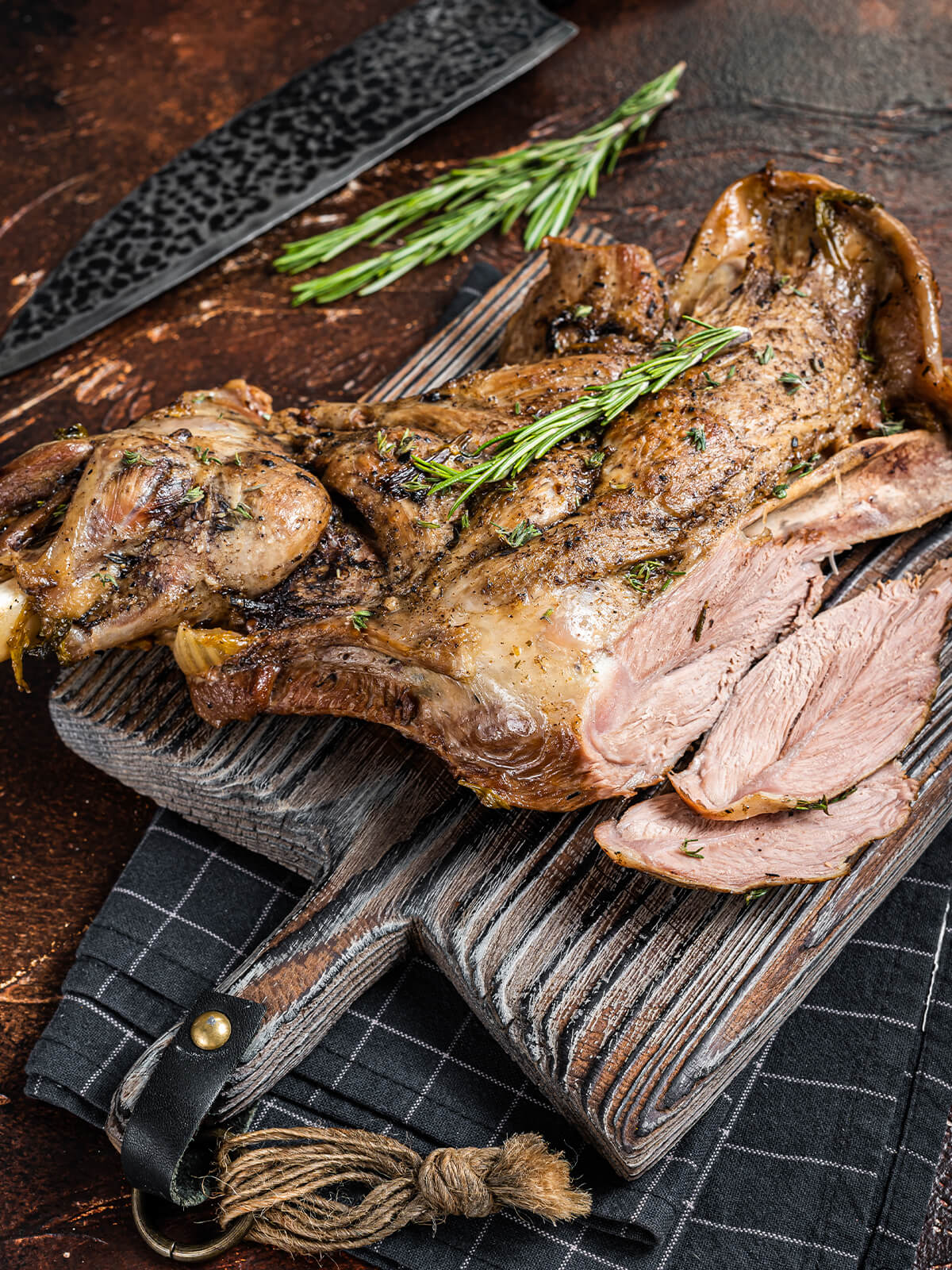 Slow-Roast Lammschulter mit Rosmarin und Thymian, auf dunklem Holzbrett angerichtet mit bereits aufgeschnittenen butterzarten Scheiben