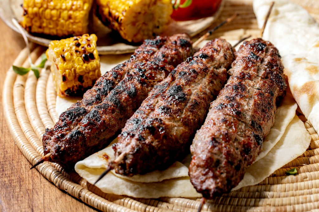 Drei goldgrillgestreifte Lyulya Kebab Spieße auf rundem Holzbrett mit gegrilltem Mais, Tomate und Lavash-Brot