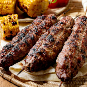 Drei goldgrillgestreifte Lyulya Kebab Spieße auf rundem Holzbrett mit gegrilltem Mais, Tomate und Lavash-Brot