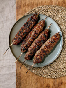 Aserbaidschanische Lyulya Kebab Hackfleisch-Spieße vom Grill mit goldbraunen Grillstreifen auf Keramikteller mit gerösteten Gemüsen