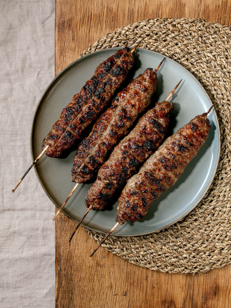 Aserbaidschanische Lyulya Kebab Hackfleisch-Spieße vom Grill mit goldbraunen Grillstreifen auf Keramikteller mit gerösteten Gemüsen