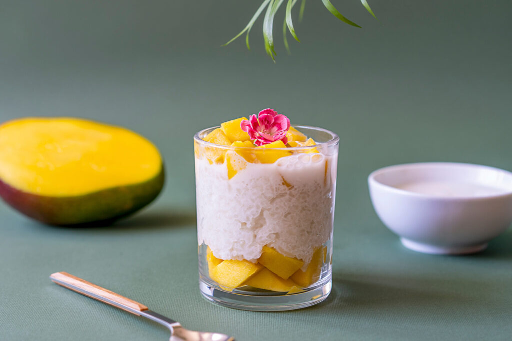 Mango Sticky Rice in Glas mit weißem Klebreis, gelben Mango-Würfeln, Kokosmilch und Edelblüte auf grünem Hintergrund
