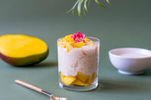 Mango Sticky Rice in Glas mit weißem Klebreis, gelben Mango-Würfeln, Kokosmilch und Edelblüte auf grünem Hintergrund