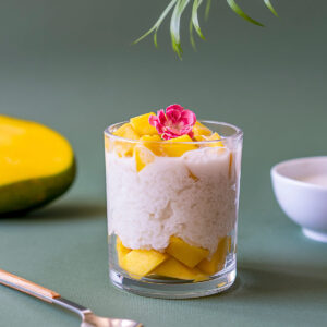 Mango Sticky Rice in Glas mit weißem Klebreis, gelben Mango-Würfeln, Kokosmilch und Edelblüte auf grünem Hintergrund