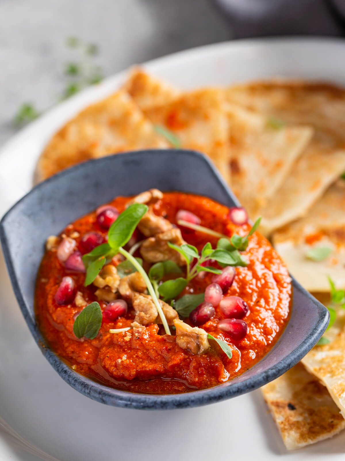 Original syrische Muhammara aus gerösteter roter Paprika und Walnüssen in blauer Keramikschale, garniert mit Granatapfelkernen, gehackten Walnüssen und frischen Oreganoblättern, dazu pfannengeröstetes Fladenbrot