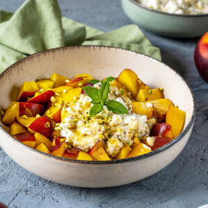 Nektarinen-Ricotta-Salat in heller Keramikschale mit gelb-roten Fruchtwürfeln, Ricotta-Mascarpone-Creme und gehackten Pistazien