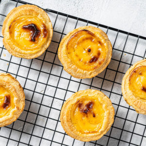 Fünf Pastéis de Nata mit dunklen Flecken auf einem schwarzen Abkühlgitter von oben