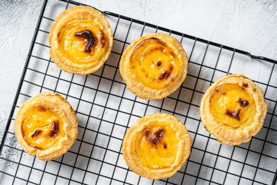 Fünf Pastéis de Nata mit dunklen Flecken auf einem schwarzen Abkühlgitter von oben